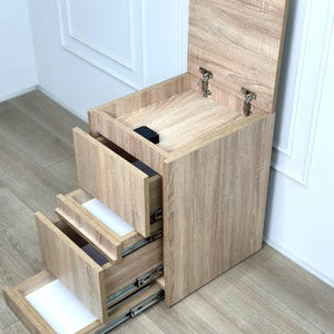 May include: A light brown wood-grain storage unit featuring a vertical stack of three pull-out drawers with metal glides and a top compartment with a hinged lid. The unit has a rectangular frame and a light oak-colored finish throughout.