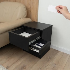 Pode incluir: Uma mesa de cabeceira preta com duas gavetas, uma aberta para revelar um interior branco. A mesa de cabeceira está ao lado de um sofá bege. Uma mão segura um cartão branco acima da mesa de cabeceira.