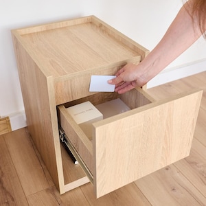 May include: A light oak wood bedside table with a single open drawer. The drawer contains a white box with the text Michael Kors printed on the top. A plain white rectangular card is held against the front panel of the drawer unit.