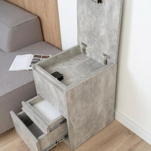May include: A grey bedside table with a concrete-like texture, featuring a top compartment with a hinged lid and two lower drawers with metal glides. The unit has a vertical orientation and is constructed with a light grey finish throughout.