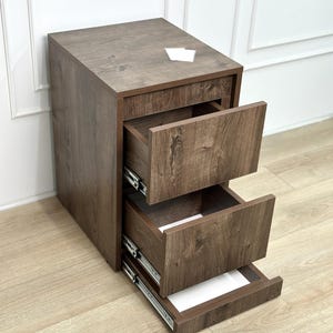 May include: A dark brown wood-grain finish filing cabinet featuring three stacked drawers with metal sliding tracks. The top surface is flat and rectangular, and the drawers are shown in an open position, revealing the interior structure.