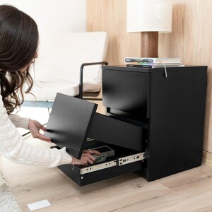 Könnte beinhalten: Ein schwarzer Aktenschrank mit zwei Schubladen und matter Oberfläche. Die obere Schublade ist geschlossen, die untere ist geöffnet und gibt den Blick auf die seitlichen Metallschienen frei. Das Möbelstück ist rechteckig und hat eine schwarze Oberfläche.