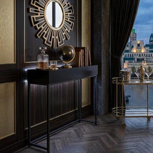 May include: A black console table featuring a glass decanter, a small glass, a globe, and three books. Above the table hangs a round sunburst mirror with gold accents. To the right, a gold metal bar cart holds a metal ice bucket and two glasses.