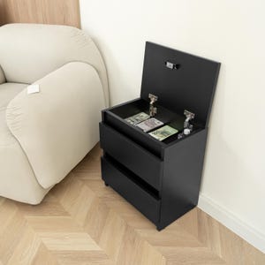 May include: A black bedside table with two drawers and a lift-up top. The top is open, displaying banknotes. The table is next to a beige armchair and a white wall. The wooden floor has a herringbone pattern.