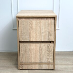 May include: A light oak wood grain bedside table featuring a rectangular top surface and two stacked drawers with a matching wood finish. The unit has a clean, vertical design with a flat base and a consistent light brown color throughout the structure.
