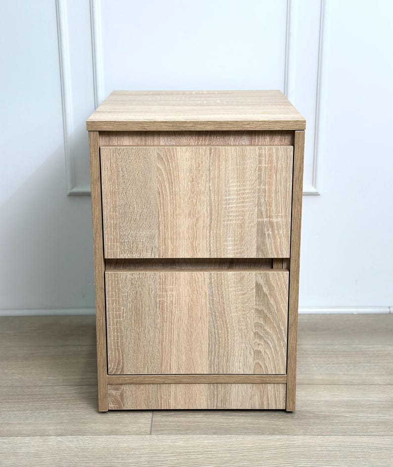 May include: A light oak wood grain bedside table featuring a rectangular top surface and two stacked drawers with a matching wood finish. The unit has a clean, vertical design with a flat base and a consistent light brown color throughout the structure.