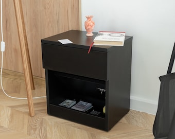May include: A black nightstand with a drawer and open shelf. A small, peach-colored vase and a book with a red bookmark sit on top. The shelf contains what appears to be currency. The nightstand is placed on a wooden floor.