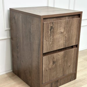 May include: A rectangular wooden filing cabinet featuring a dark brown wood grain finish. The unit is constructed with a flat top surface and two stacked drawers, each equipped with a horizontal recessed handle along the top edge of the drawer front.