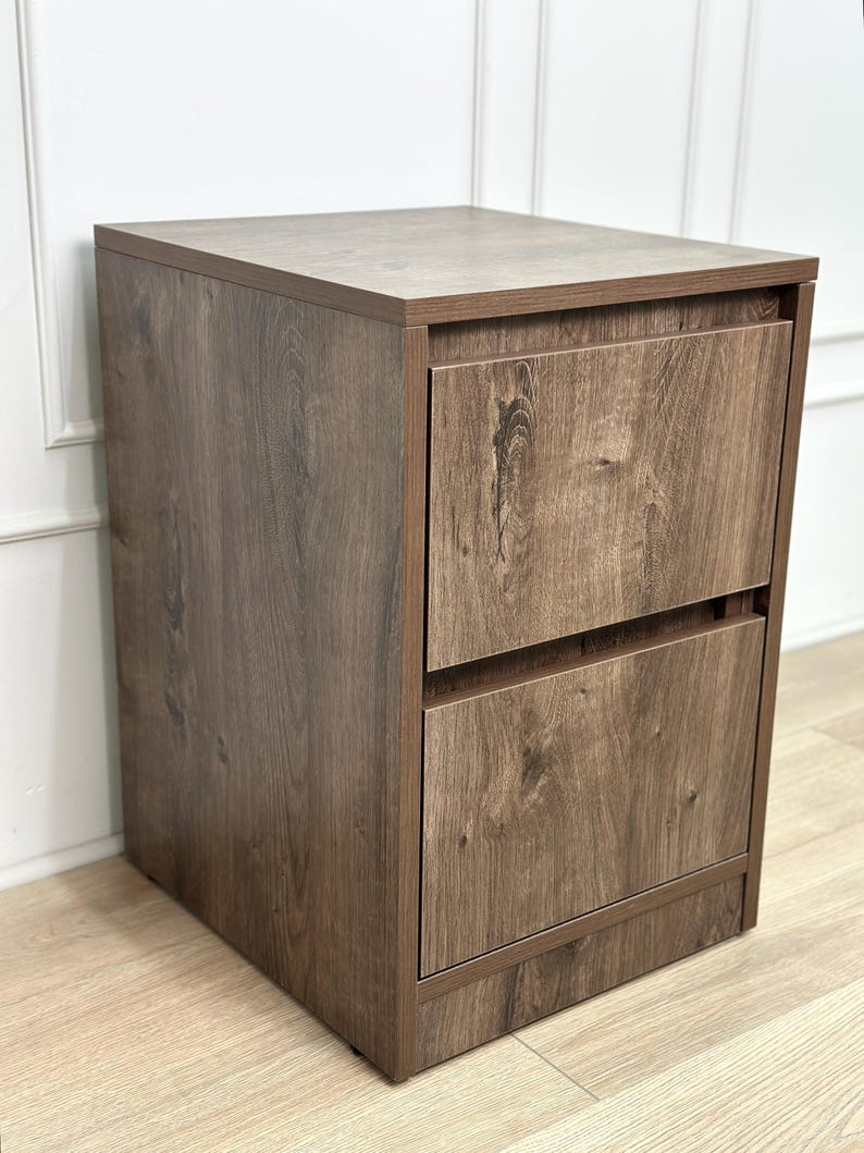 May include: A rectangular wooden filing cabinet featuring a dark brown wood grain finish. The unit is constructed with a flat top surface and two stacked drawers, each equipped with a horizontal recessed handle along the top edge of the drawer front.