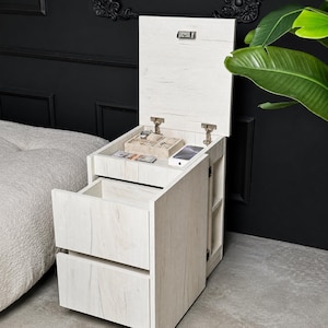 May include: A light wood grain bedside table featuring two pull-out drawers and a top compartment with a hinged lid. The interior of the top section contains a beige jewellery organiser tray and a white smartphone. The unit has a vertical side shelf.
