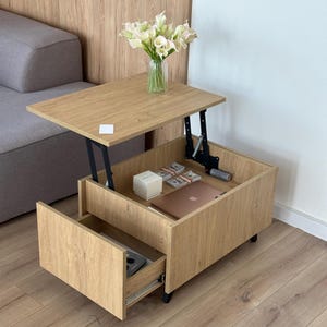 Peut inclure: Une table basse en bois de chêne clair avec un mécanisme de plateau relevable, un tiroir coulissant inférieur et un compartiment de rangement ouvert contenant une boîte blanche, des liasses de billets et un ordinateur portable.
