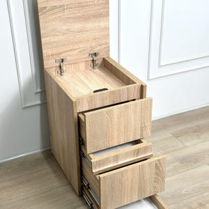 Könnte beinhalten: Ein helles Holzaufbewahrungsmöbel mit einem hochklappbaren Deckel und drei ausziehbaren Schubladen. Der Schrank hat eine Holzmaserung und Metallbeschläge. Der obere Deckel ist geöffnet.