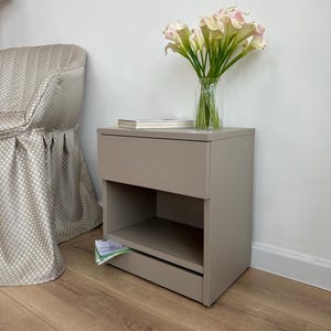 May include: A taupe-colored nightstand with a drawer and open shelf. A clear glass vase holds calla lilies with pink tips. A book rests on top. The nightstand is next to a patterned armchair.