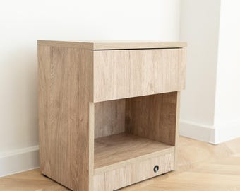 May include: A light oak nightstand with a drawer and an open shelf. The nightstand has a rectangular shape and a simple, modern design. The wood grain is visible, giving it a natural look. The nightstand is placed against a white wall.