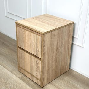 May include: A light brown wood grain bedside table featuring a square top surface and two stacked drawers with a flat front design. The unit has a clean, boxy silhouette with a consistent light oak-colored finish throughout the entire structure.