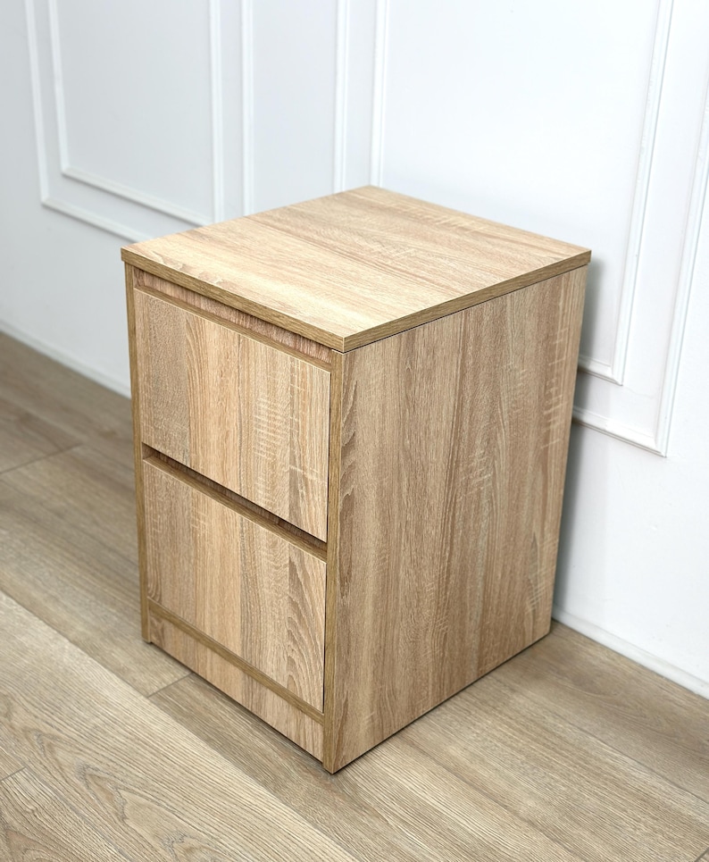 May include: A light brown wood grain bedside table featuring a square top surface and two stacked drawers with a flat front design. The unit has a clean, boxy silhouette with a consistent light oak-colored finish throughout the entire structure.