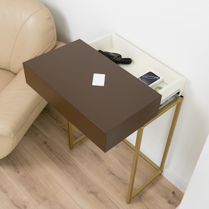 Puede incluir: Una mesa auxiliar moderna con una tapa rectangular marrón y un cajón interior blanco. El cajón está parcialmente abierto, revelando un guante negro, un teléfono inteligente y otros objetos. La mesa tiene una estructura metálica dorada.