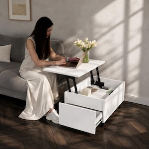 May include: A white rectangular coffee table with a lift-top mechanism that elevates a portion of the surface for use as a desk. The table features a lower storage compartment and a pull-out drawer, with a glass vase of white flowers on top.