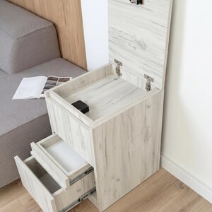 May include: A light wood grain bedside table with a vertical lift-top compartment featuring a metal latch and two pull-out drawers with metal sliding tracks. The unit has a rectangular frame and a light beige finish with visible wood grain patterns.