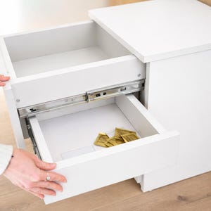 Pode incluir: Um armário branco com duas gavetas abertas. A gaveta superior está vazia, enquanto a gaveta inferior contém vários pacotes dourados. O armário está sobre um chão de madeira clara.