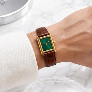 Pode incluir: Um relógio de pulso retangular com caixa dourada, mostrador verde esmeralda vibrante com ponteiros dourados e pulseira de couro marrom com padrão de crocodilo texturizado. O relógio possui uma pequena coroa dourada na lateral.