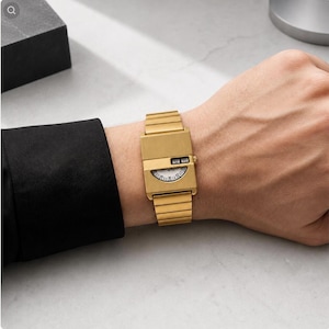Könnte beinhalten: Eine goldfarbene Armbanduhr mit rechteckigem Gehäuse und passendem Metallgliederarmband. Das Zifferblatt zeichnet sich durch ein markantes Design mit einem digitalen Zeitanzeigefenster oben und einer halbkreisförmigen Minutenskala unten aus.