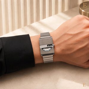 May include: A brushed silver-tone metal wristwatch with a rectangular case and a matching link bracelet. The watch face features a unique horizontal digital display window at the top and a semi-circular analogue dial below, showing time markers.