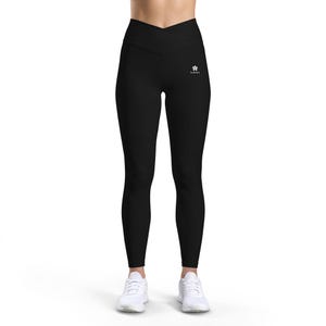 Peut inclure: Legging noir uni doté d'une taille haute avec un design croisé en forme de V sur le devant. La hanche gauche présente un petit logo graphique blanc composé d'une icône de fleur stylisée au-dessus du texte LAIVI en police sans empattement.