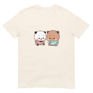 May include: A cream-colored short-sleeve t-shirt featuring a central graphic of two cartoon bears. One bear is white with black ears and a pink shirt, while the other is brown with a light blue shirt. Both characters have round faces and rosy cheeks.