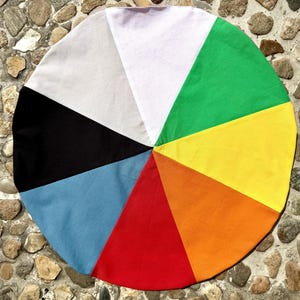 Könnte beinhalten: Ein kreisförmiges Stoffstück, das aus acht verschiedenen dreieckigen Segmenten in einer lebendigen Farbkreis-Anordnung besteht. Die Farben umfassen Weiß, Grün, Gelb, Orange, Rot, Hellblau, Schwarz und Hellgrau, die alle im Mittelpunkt zusammenlaufen.