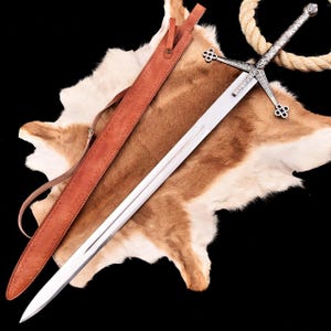 May include: A long, silver sword with an ornate hilt and crossguard, resting on a brown and white hide. A brown leather sheath with a strap lies beside the sword. The sword's blade is approximately 36 inches long.