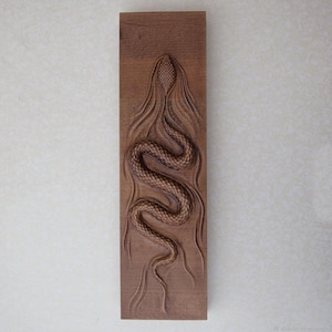 May include: A rectangular wooden carving of a snake in relief. The snake is shown in a winding pose, with textured scales and a detailed head. The wood has a warm brown tone, and the carving is set against a neutral background.