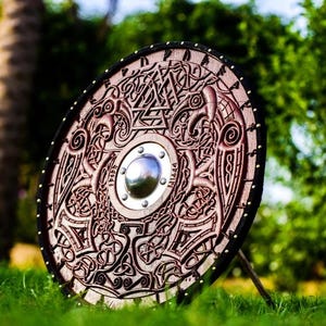 May include: A round wooden shield with intricate carvings of a bird and knotwork patterns. The shield is painted a dark brown color and has a metal center boss. The shield is standing on a green grassy surface.