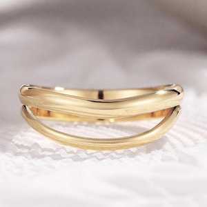 May include: A gold-colored ring with a double-band design. The top band has a wavy, organic shape, while the lower band curves smoothly. The ring is set against a white, textured background, with soft lighting.