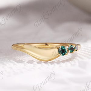 Könnte beinhalten: Ein goldener Ring mit fließendem Design, der drei kleine, runde, teal-farbene Edelsteine ​​aufweist. Der Ring hat eine glatte, polierte Oberfläche und ist vor einem weichen, weißen Hintergrund platziert.