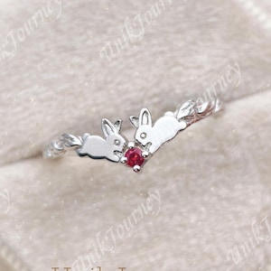 May include: A silver-toned ring featuring two rabbit silhouettes facing each other, with a small, round, vibrant red gemstone set between them. The band has a textured, vine-like appearance, and the ring is presented in a soft, light-colored box.