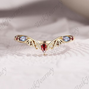 May include: A gold-colored ring with a unique design. The ring features a central red gemstone and two blue gemstones on either side. The ring's design includes wing-like patterns. The ring is set against a white background.