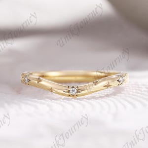 May include: A gold chevron-shaped ring featuring a polished band with engraved starburst patterns. Three small, round, clear stones are set within the center of the starburst engravings, spaced evenly along the curved surface of the band.