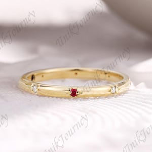 May include: A gold band ring with a small red gemstone in the center and two small white gemstones on either side. The ring has a smooth, polished finish and is set against a soft, white background.