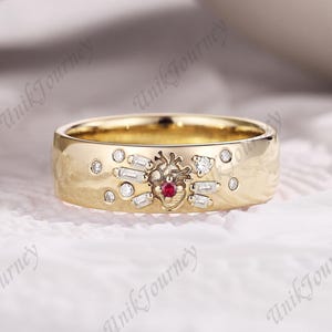 May include: A gold-colored ring with a central heart-shaped design, accented with a small red gemstone and surrounded by small, clear gemstones. The ring also features several rectangular and round clear gemstones.