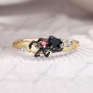 May include: A gold-toned ring featuring a textured band with a black spider motif. The spider has a small round red gemstone and a larger black heart-shaped gemstone set into its body. Small silver web accents are visible on the band.