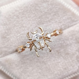 May include: A gold ring featuring a central beetle-shaped design adorned with clear baguette and round-cut stones. The insect motif has articulated legs and a faceted body, with additional small round stones set along the thin metal band.