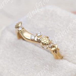 May include: A gold-toned curved ring featuring a small sea turtle motif and three clear, round-cut stones set along the band. The ring has a polished finish and a delicate, contoured shape that follows a gentle wave-like arc across the finger.