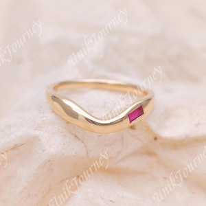 May include: A gold band ring with a curved, wave-like silhouette featuring a single, small, rectangular-cut red gemstone set flush into the metal surface. The ring has a polished, smooth finish and a simple, continuous design.