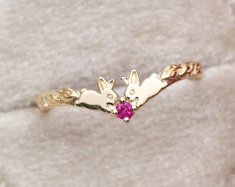 Könnte beinhalten: Ein goldener V-förmiger Ring mit zwei kleinen Hasenfiguren, die sich in der Mitte gegenüberstehen. Ein einzelner, runder, leuchtend pinker Edelstein ist zwischen den beiden Hasen gefasst. Die Ringschiene hat ein strukturiertes Rankenmuster.