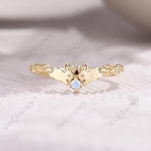 May include: A gold ring featuring two facing rabbit figures at the center, joined by a small, round, iridescent moonstone. The band displays a textured, vine-like pattern extending from the rabbits toward the sides of the ring.