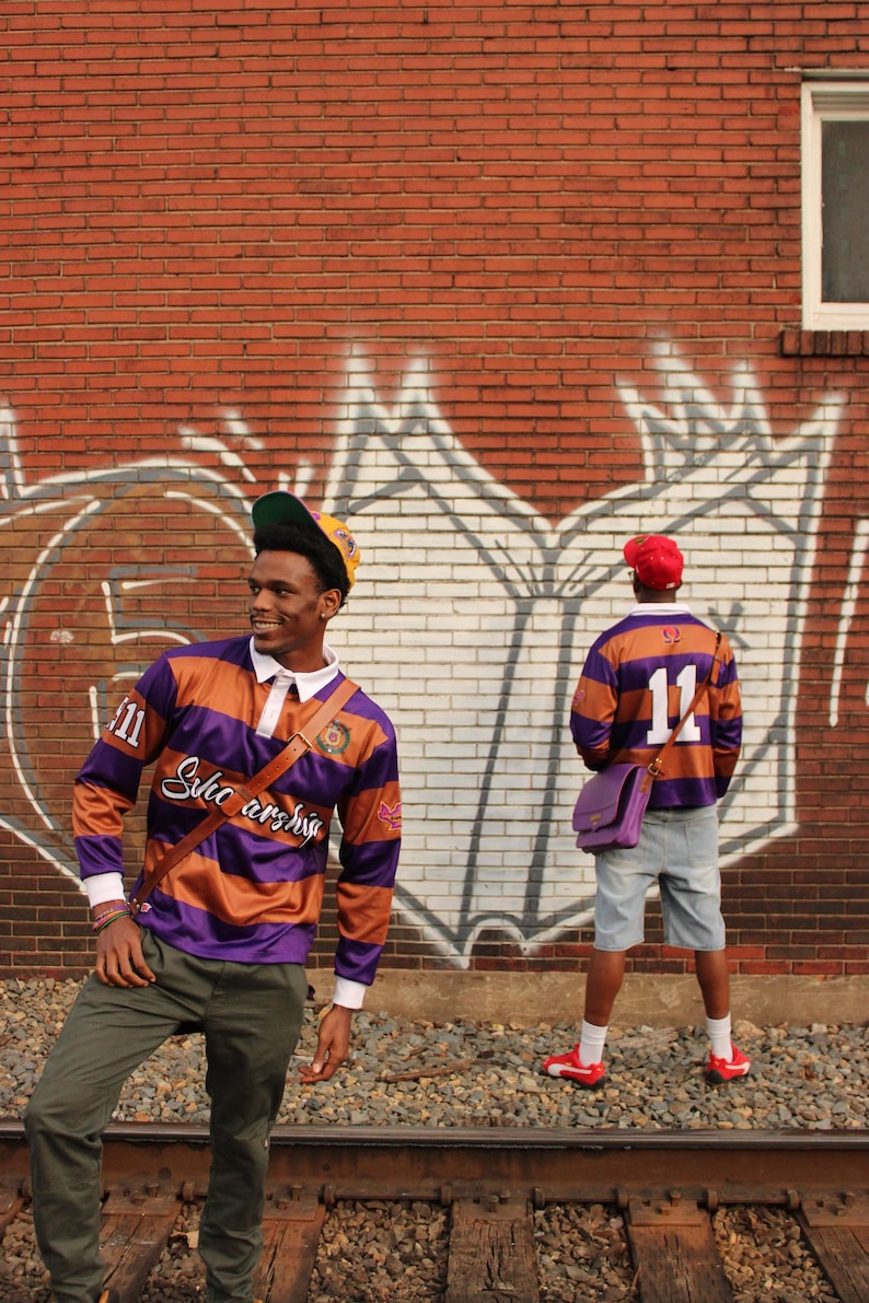 May include: Two people wearing purple and orange striped rugby shirts with the number 11. The shirts have white collars and the text "Solic archis" on the front. One person wears a yellow and green cap, the other a red cap.
