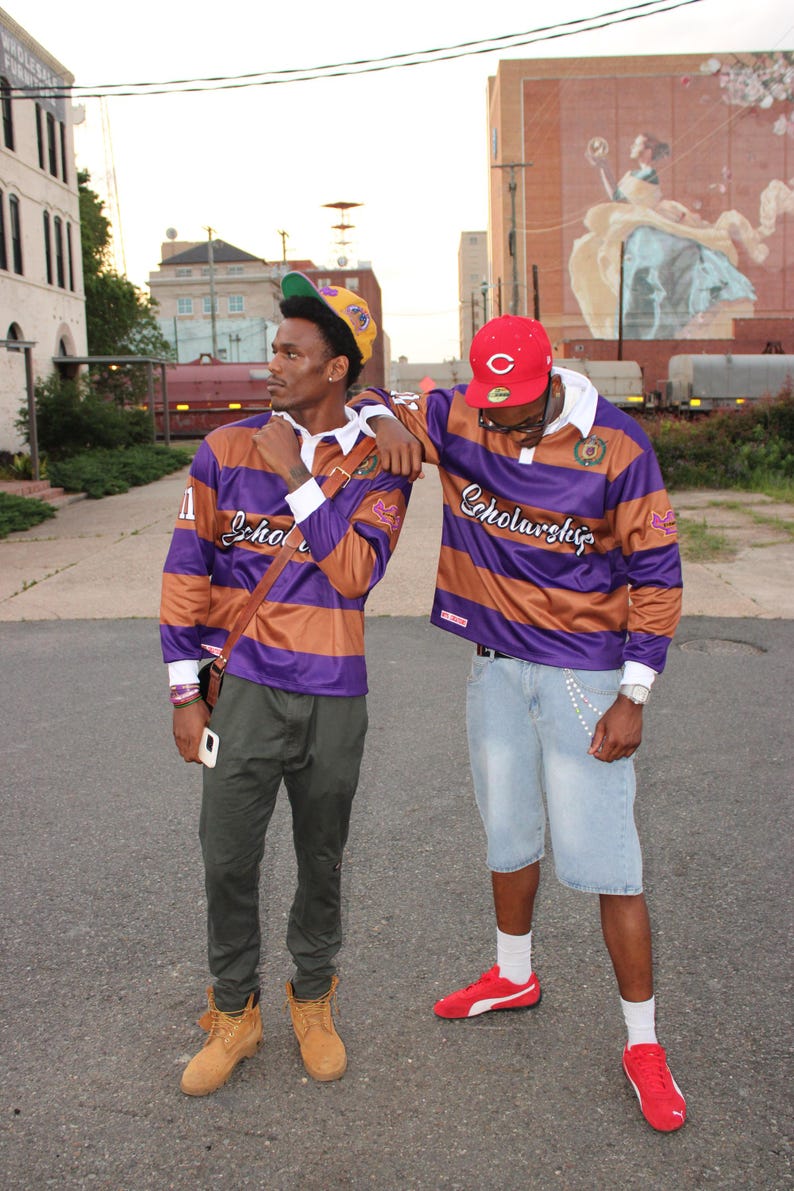May include: Two people wearing purple and brown striped rugby shirts with the word "Scholarship" on them. One person wears a yellow and green baseball cap, the other a red baseball cap. They are standing on a paved road.