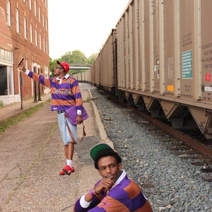 May include: Two people wearing purple and orange striped rugby shirts and red hats pose near a train. One person is taking a photo with a phone, while the other is sitting on the ground. A purple bag is visible.