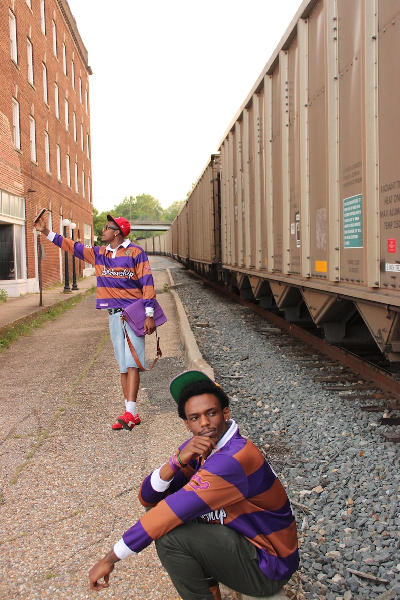 May include: Two people wearing purple and orange striped rugby shirts and red hats pose near a train. One person is taking a photo with a phone, while the other is sitting on the ground. A purple bag is visible.